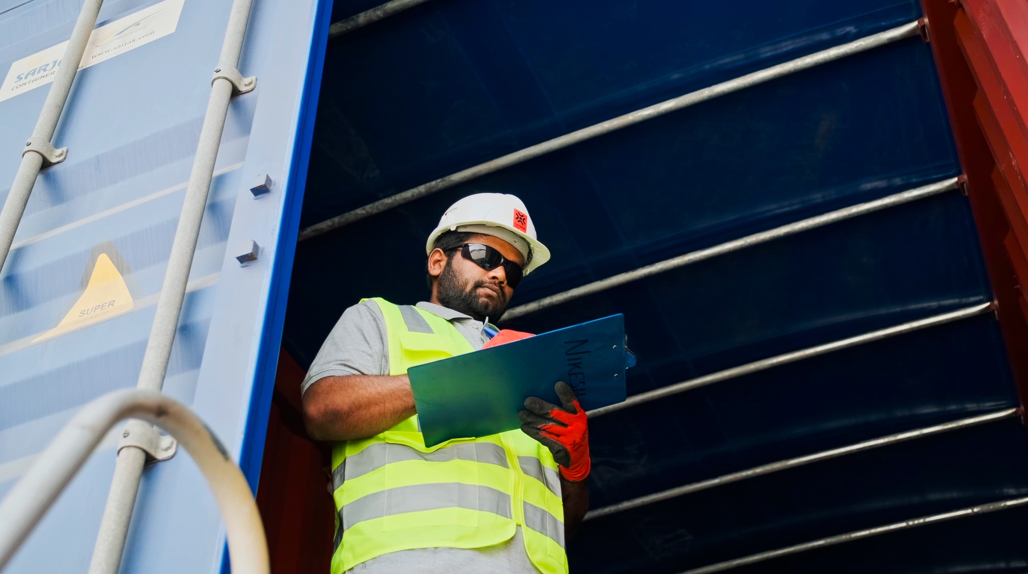 A construction worker in a safety vest and hard hat stands inside a shipping container, holding a clipboard. He appears focused, with a serious expression. 
