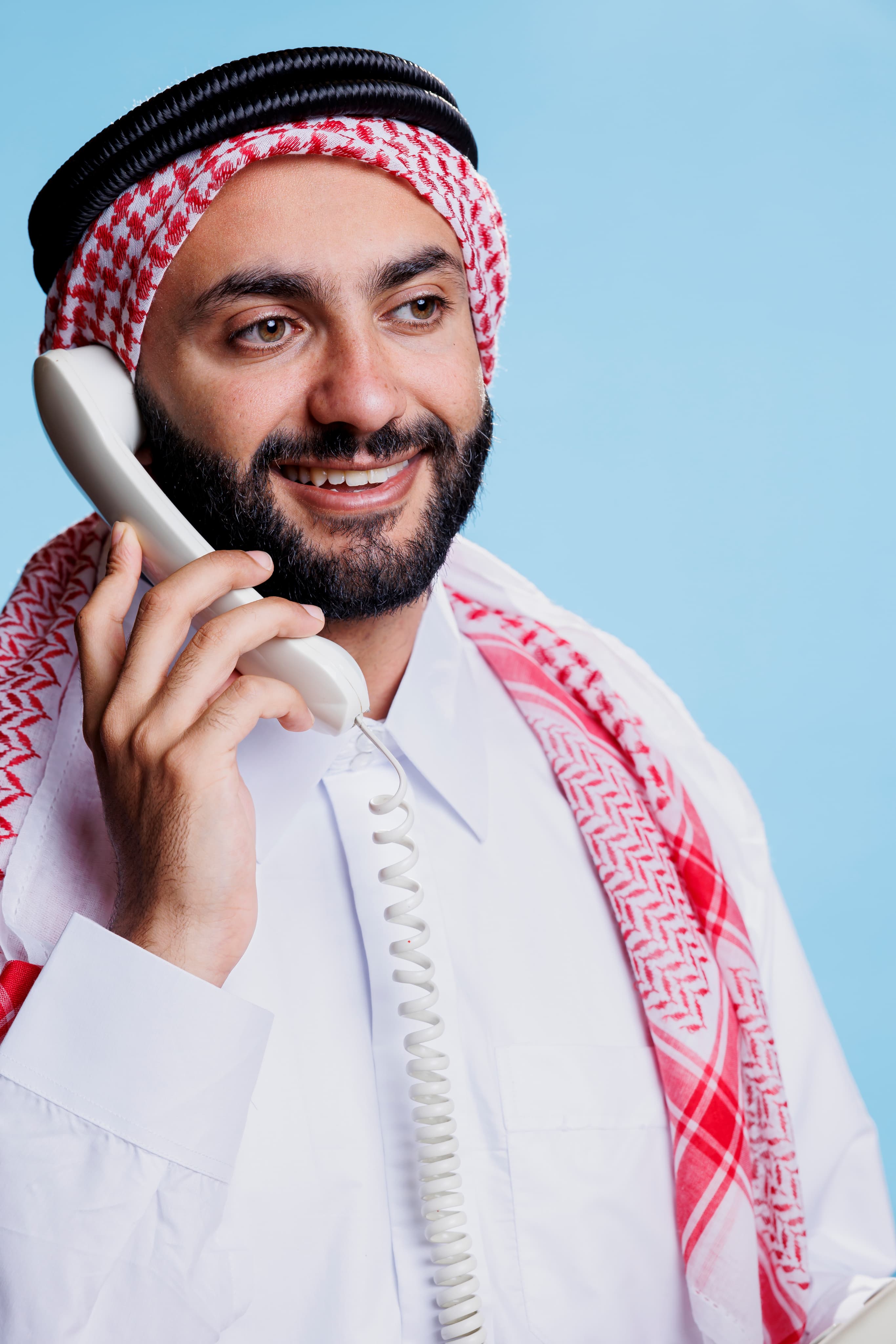 A person in traditional Arab dress holds a vintage telephone to their ear, standing against a light blue background. 