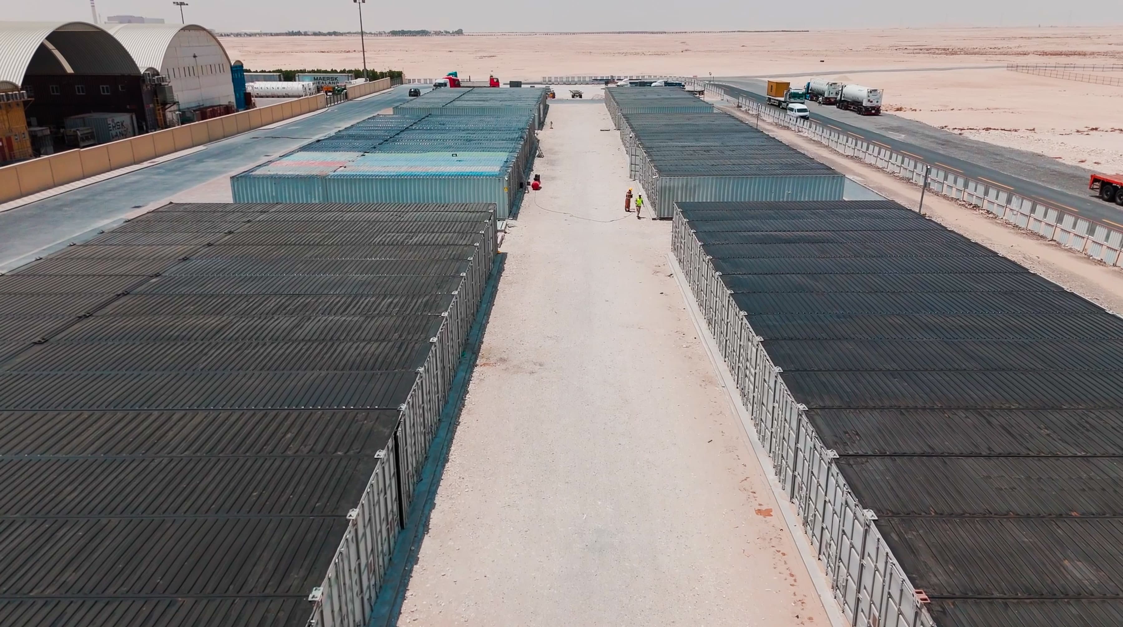Aerial view of a desert shipping container yard with rows of containers and trucks, flanked by light structures and unpaved pathways.
