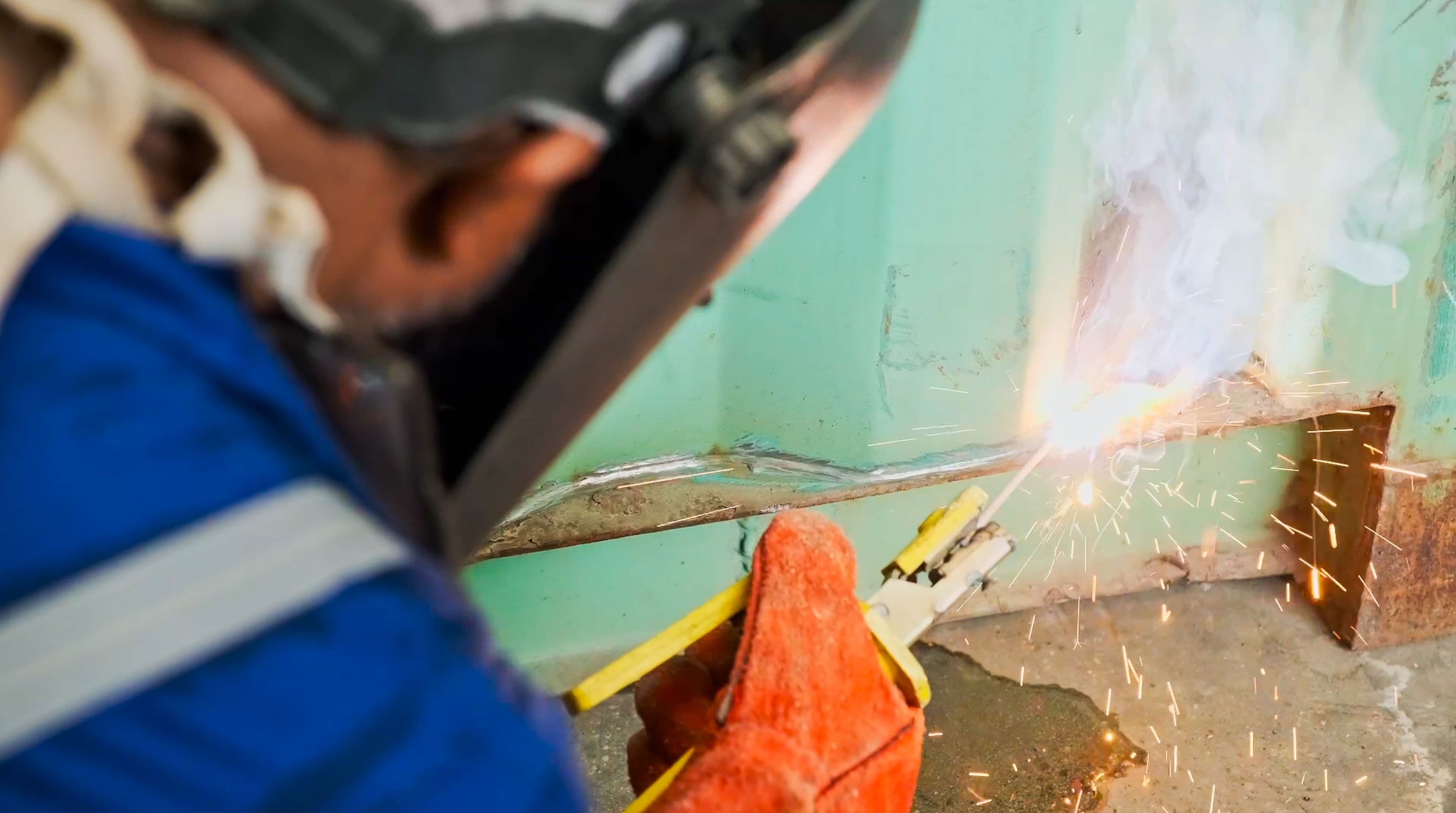 A welder, wearing a protective helmet and red gloves, uses a welding tool, creating bright sparks and smoke against a metal surface. The focus is on precision. 