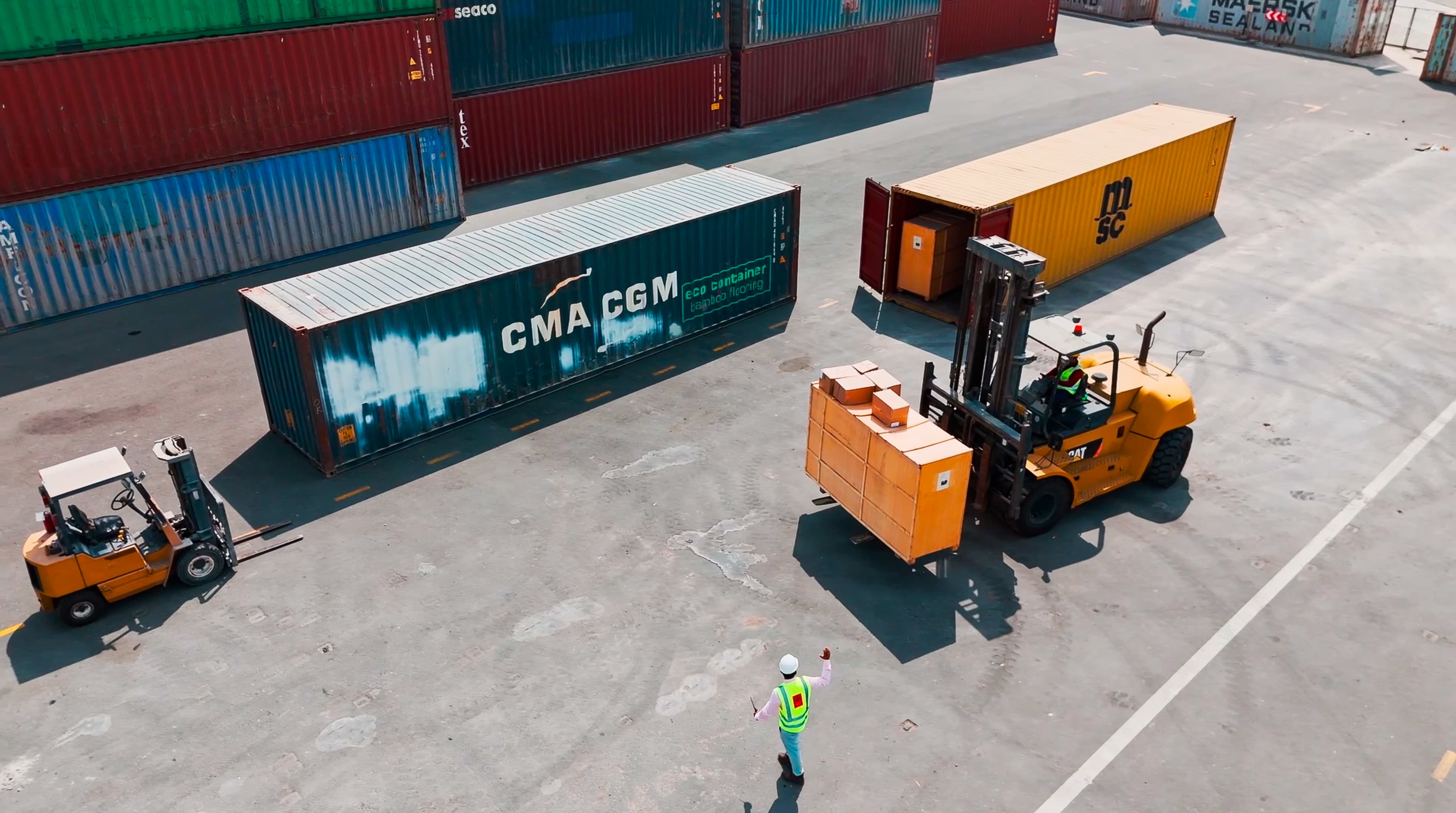 An aerial view of a logistics yard shows two forklifts moving large containers between colorfully stacked shipping containers. A worker in a reflective vest guides them, highlighting coordination and industrial efficiency.