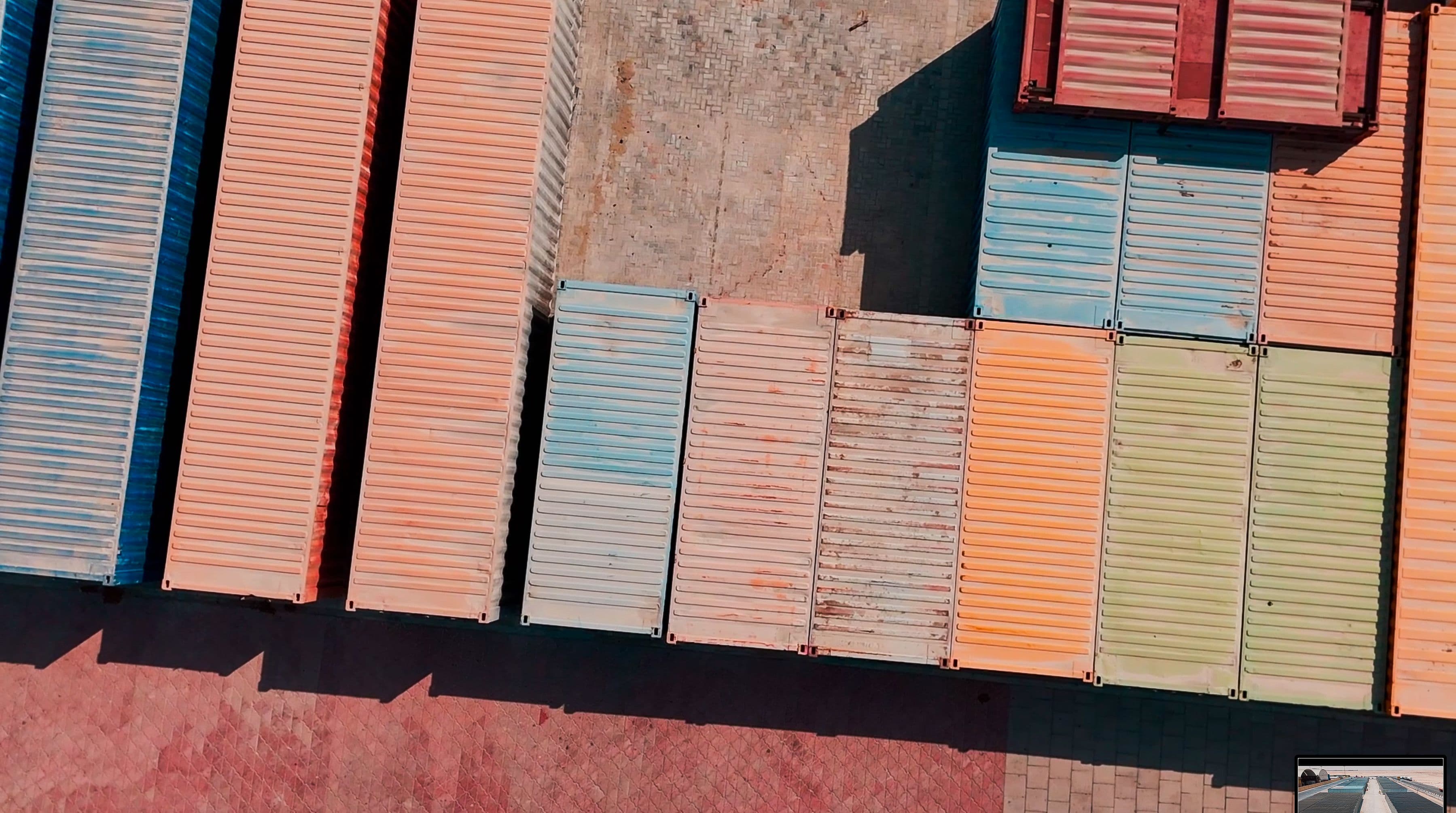 Aerial view of variously colored shipping containers in neat rows on a concrete surface, casting long shadows. The tone is orderly and industrial. 