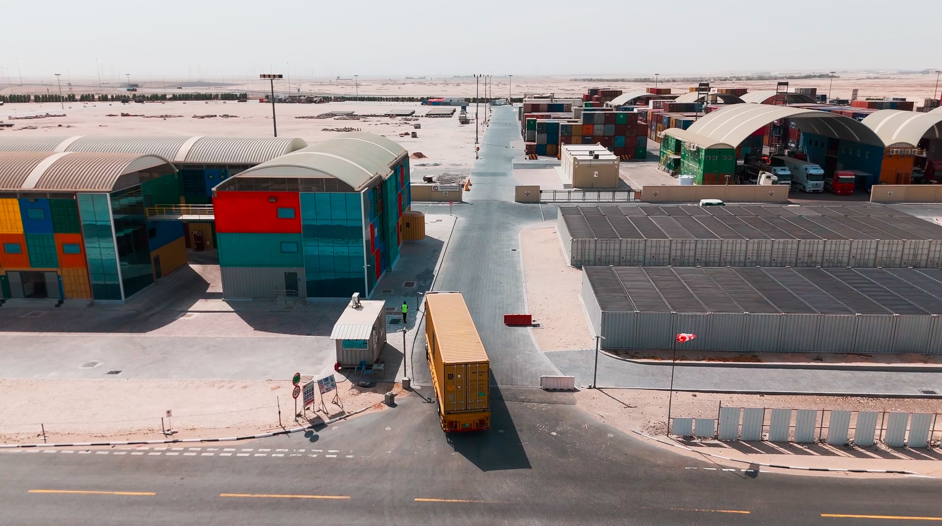Aerial view of a vibrant industrial area with colorful buildings, storage containers, and a large truck on a road. The landscape is arid and expansive