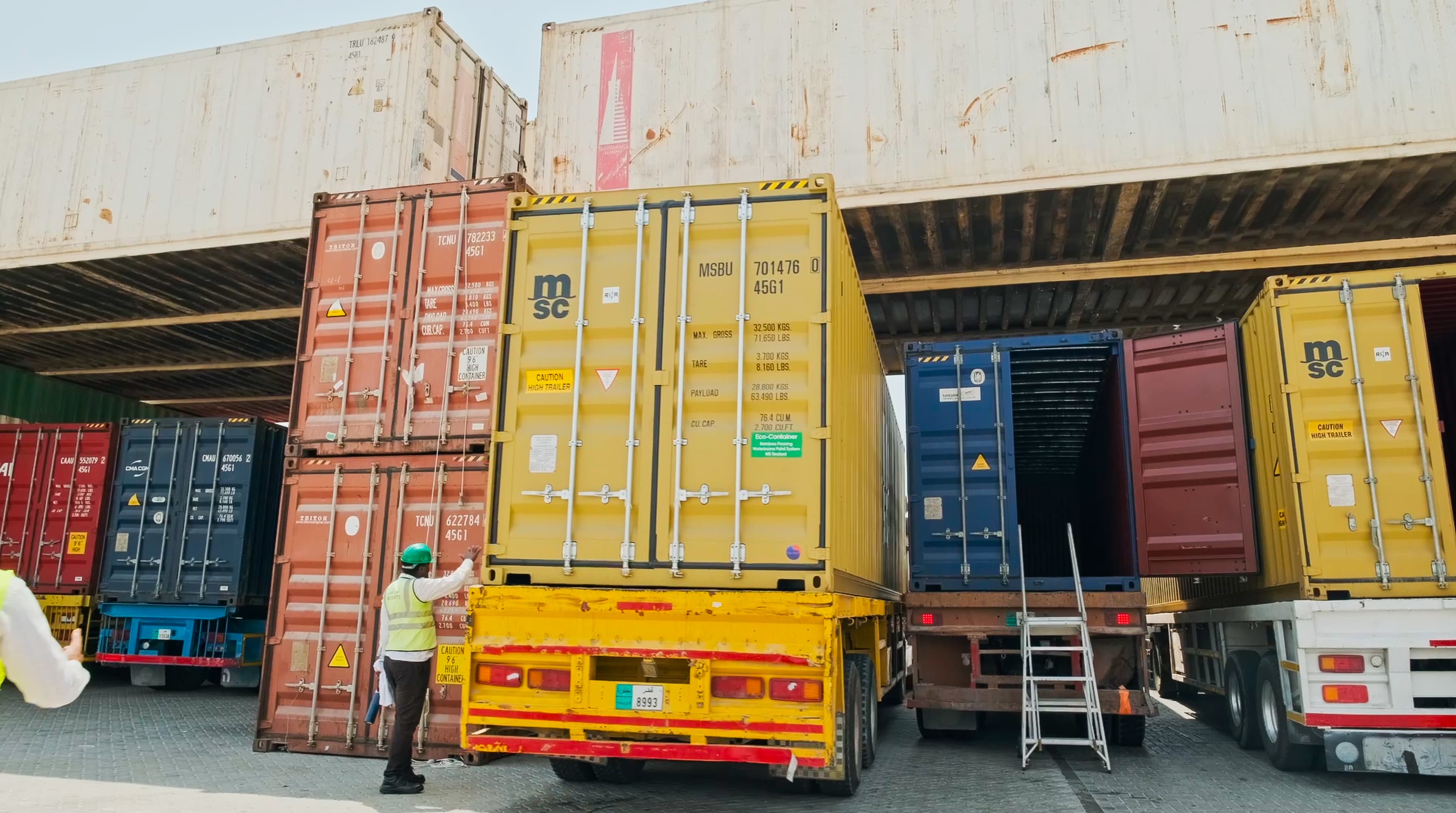 A worker in a yellow vest and hard hat opens a yellow shipping container on a truck at a busy cargo area. Containers are stacked in various colors.