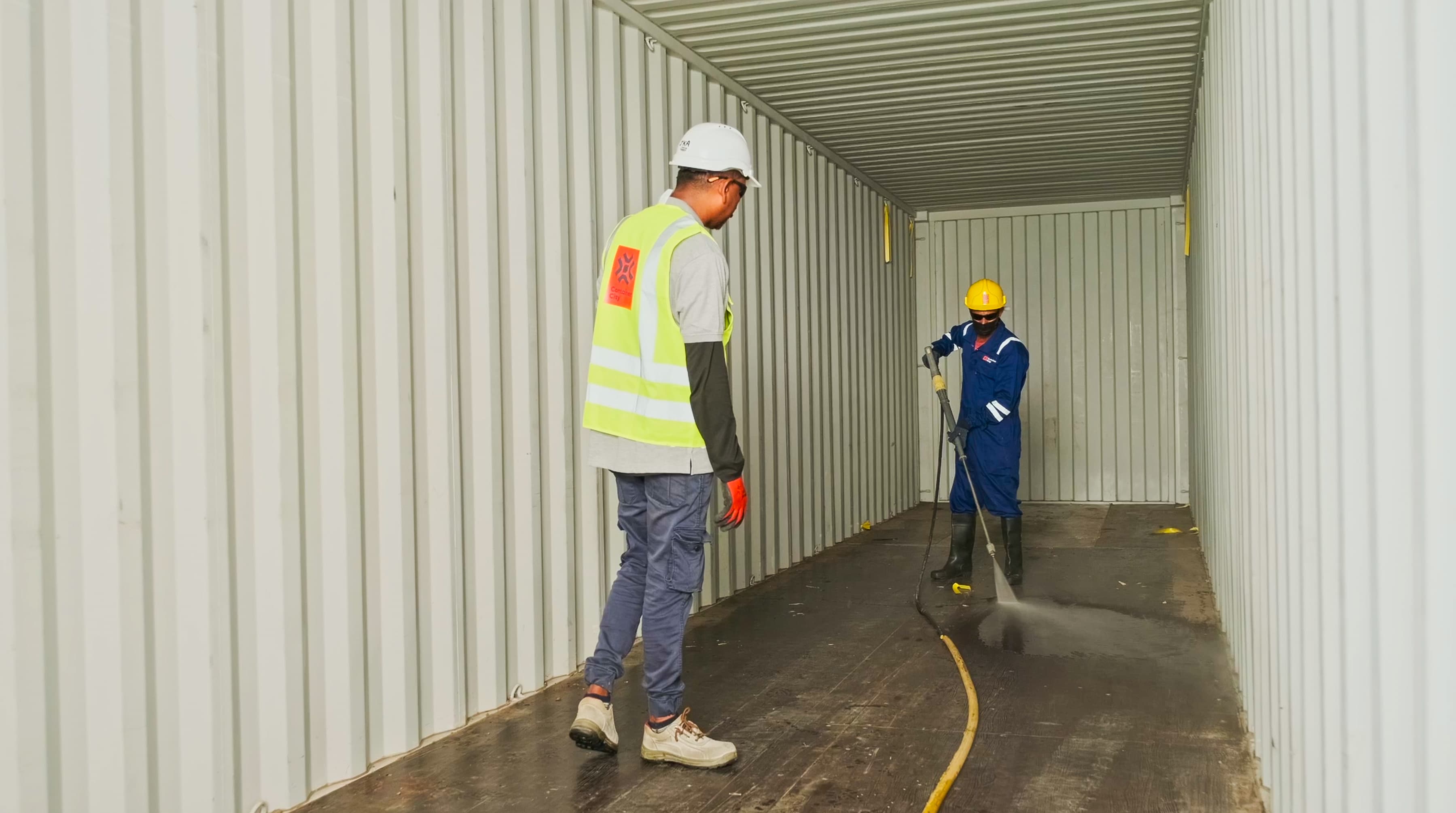Two workers in protective gear clean inside an empty shipping container. One operates a pressure washer, while the other observes, ensuring safety.