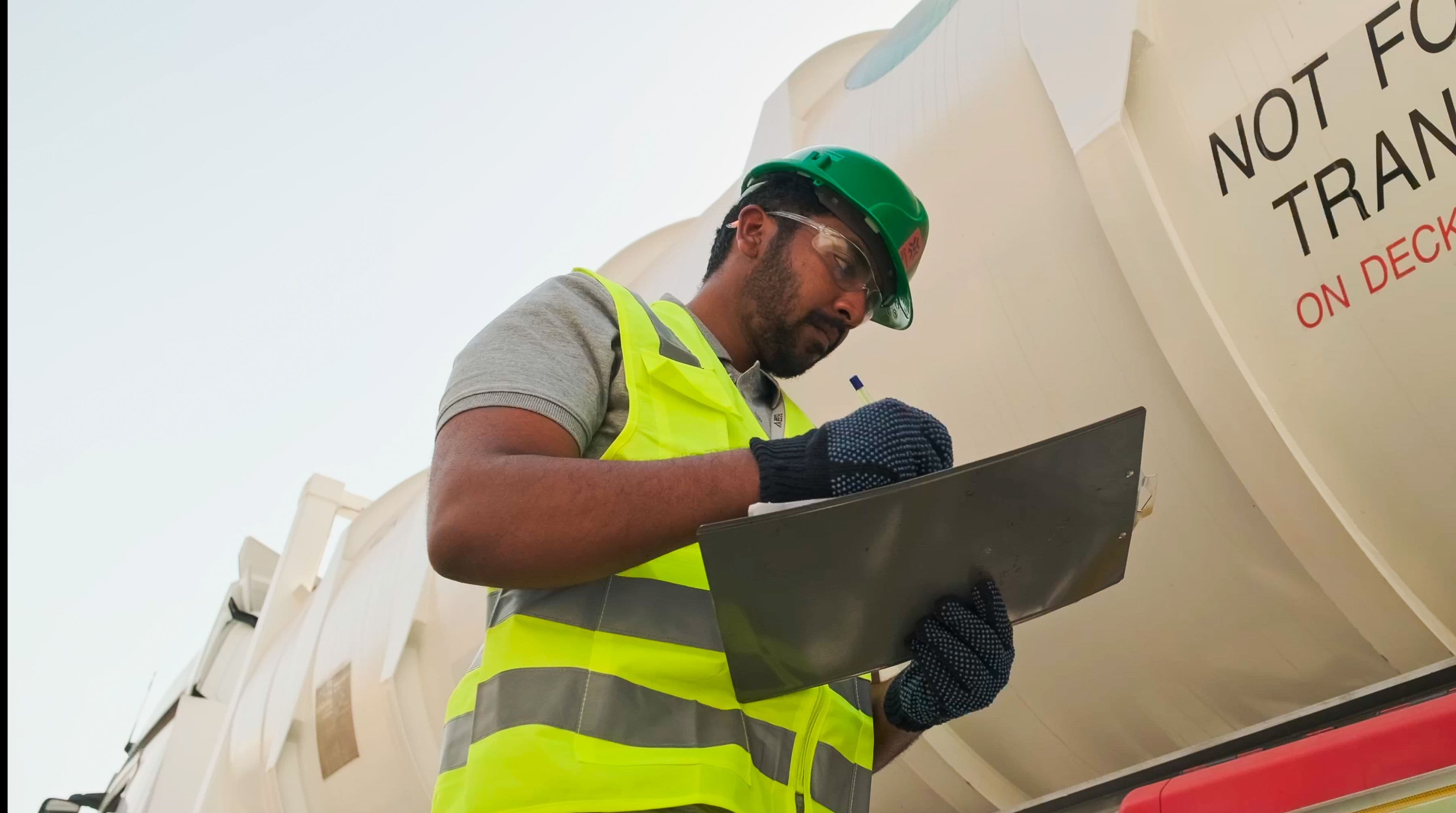A person in a hard hat and safety vest writes on a clipboard in front of a large tank with warning text. The scene conveys a sense of caution and responsibility.