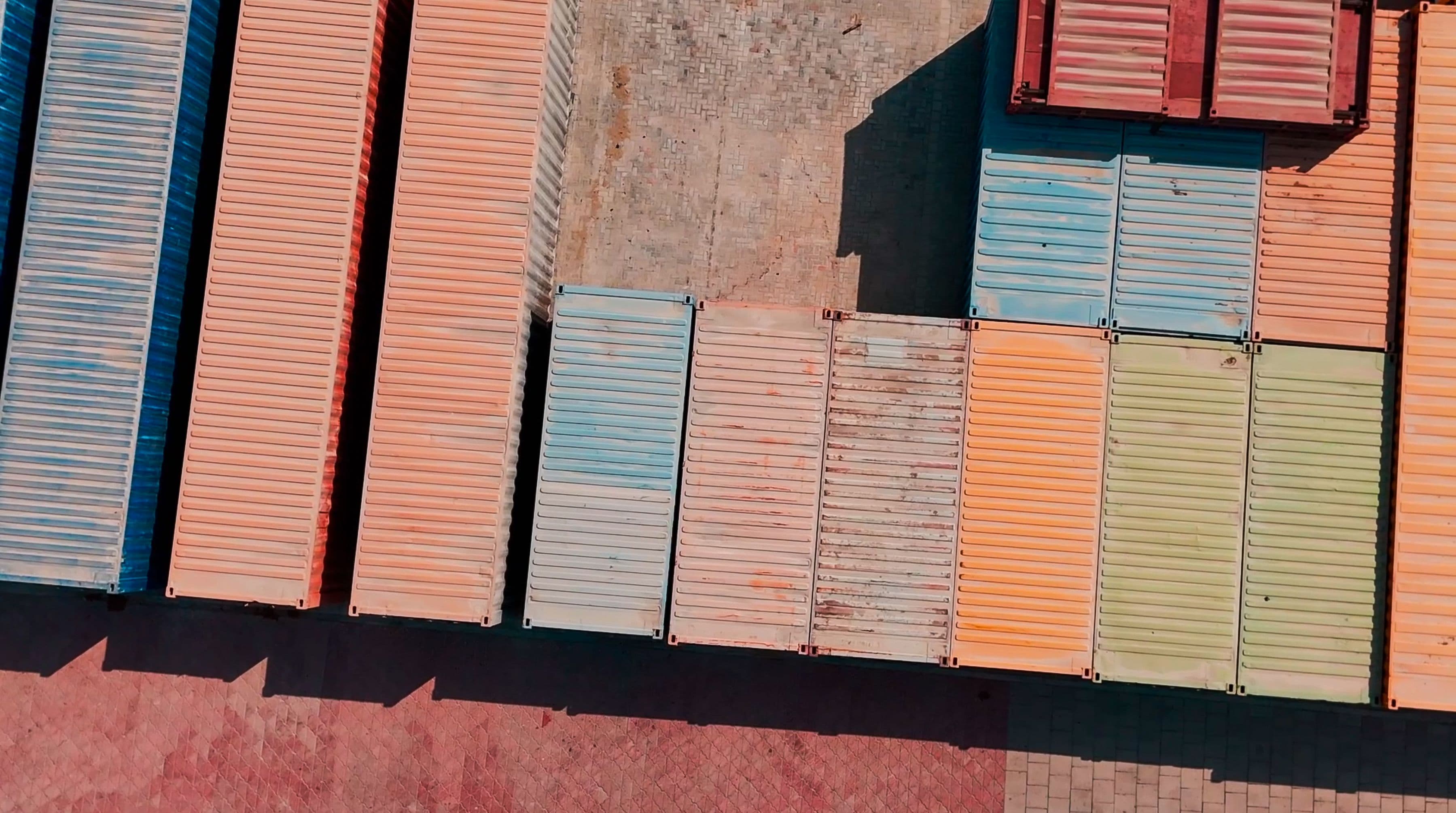 Aerial view of colorful shipping containers lined up on a concrete dock. The containers are blue, red, and orange, creating a vibrant geometric pattern.