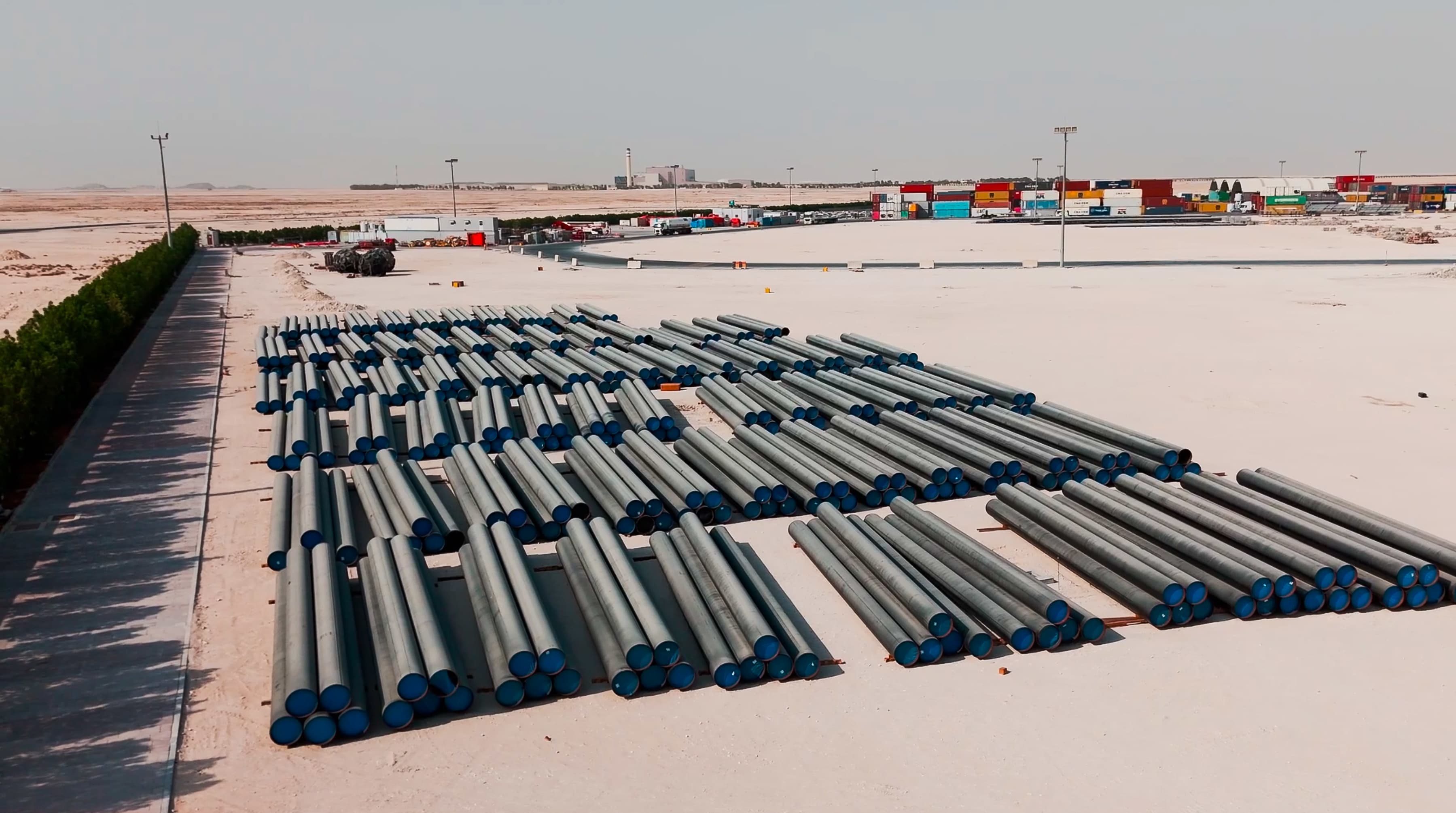 An array of large metal pipes with blue ends is neatly arranged on a sunlit industrial site. Shipping containers are in the background, under a clear sky.